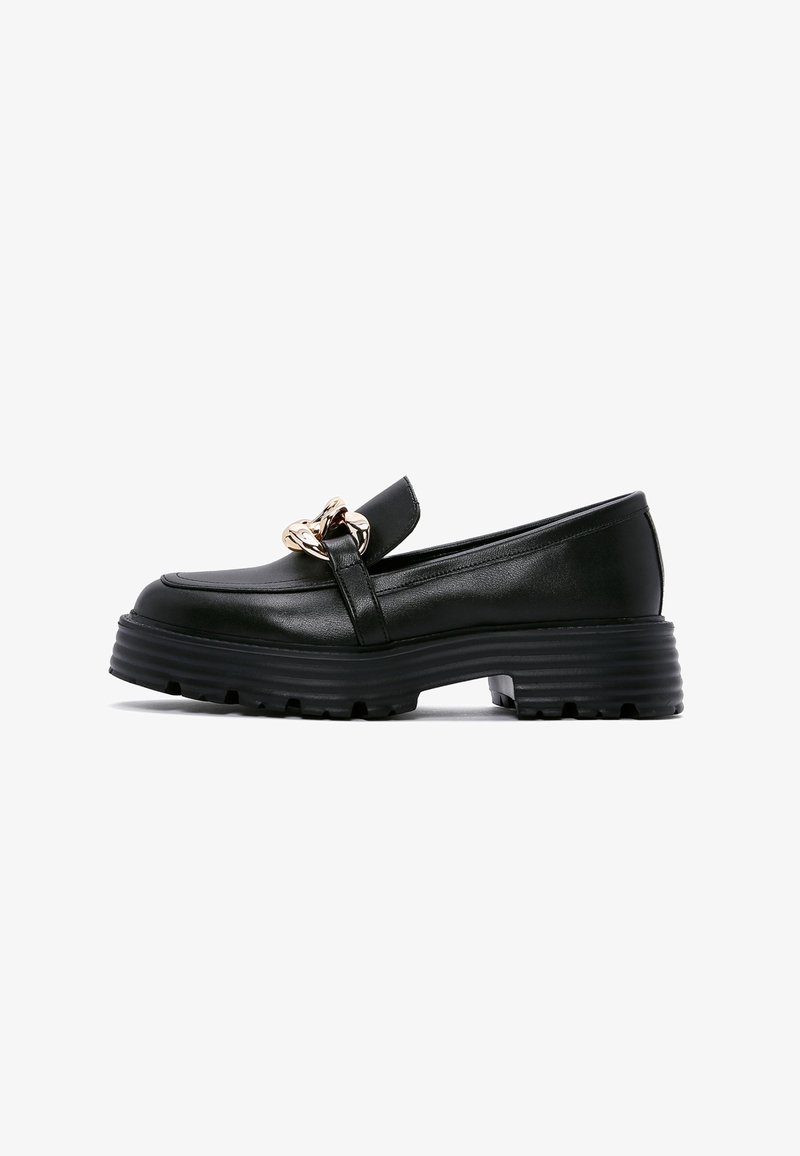 Mocassin en cuir noir avec semelle épaisse côtelée et détail de chaîne dorée sur le devant, photographié sur un fond blanc.