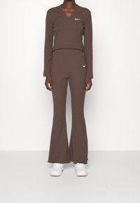 Brun ribbad långärmad topp med V-ringning och matchande utsvängda byxor. Har en vit Nike-logotyp på bröstet och midjan. Vita sneakers.