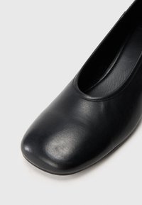 Black leather flat shoe with a rounded square toe, smooth texture, and open sides. No visible hardware or patterns.