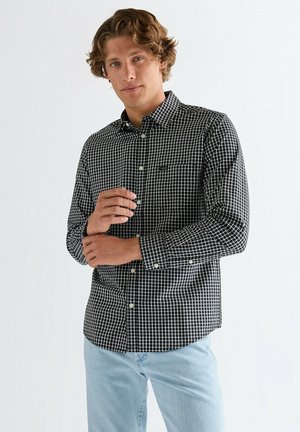 Młody mężczyzna poprawiający czarno-białą, kraciastą koszulę zapinaną na guziki, ubrany w jasnoniebieskie dżinsy, stojący na tle jednolitego białego tła.