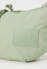 Helles grünes, weiches Stofftäschchen mit einem strukturierten Gummi-Logo-Patch und einem geknoteten Verschluss am oberen Ende. Glatte Textur, abgerundete Form.