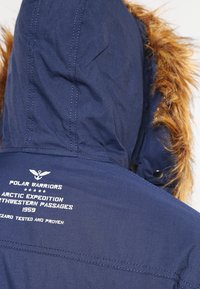 Mornarsko modra jakna s kapuco in umetnim krznom, z belimi napisi in logotipom na hrbtni strani, izdelana iz trpežnega materiala.