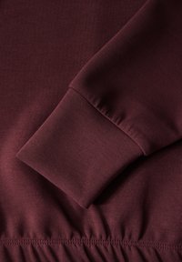 Sametová tkanina vínové mikiny s hladkým povrchem. Detail v blízkém záběru na manžetu a stažený lem, s elastickým švem na spodní části.