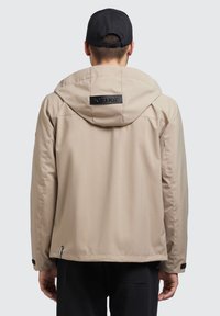 Šviesiai beige spalvos lengvas švarkas su kapišonu, turintis juodą logotipo užsegimą gale, lygia tekstūra ir reguliuojamais rankogaliais.