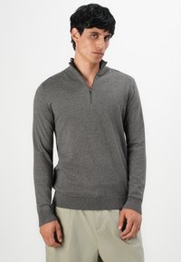 Uomo che indossa un pullover grigio a maniche lunghe con una breve cerniera al collo e pantaloni beige chiaro, in piedi davanti a uno sfondo bianco puro.
