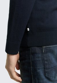 Marineblauwe gebreide trui met geribbelde boorden en zoom, voorzien van een klein wit label aan de zijkant. Gedragen over een donkere spijkerbroek.