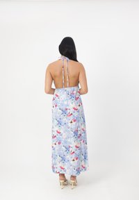 Robe maxi florale en bleu clair avec des motifs rouges et violets. Présente un col halter, un dos nu et un tissu délicat tombant jusqu'au sol.