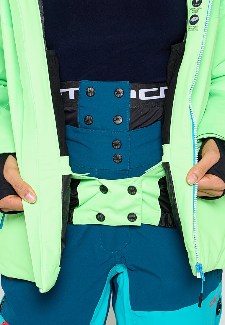 Helle grüne wasserdichte Jacke mit schwarzen und türkisfarbenen Akzenten; sichtbare Druckknöpfe und Reißverschlüsse; über einem dunklen Oberteil und türkisfarbenen Hose getragen.