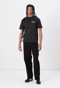 Zwart katoenen t-shirt met korte mouwen en een geel logo op de borst, gecombineerd met zwarte spijkerbroek met rechte pijpen en zwart gestreepte sneakers.
