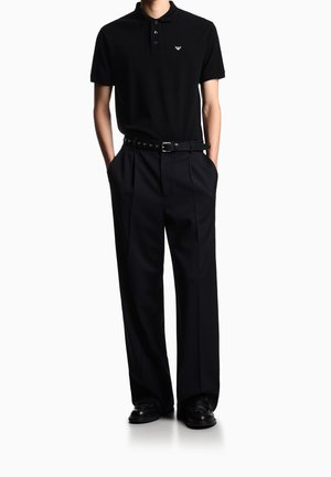 Mann trägt schwarzes kurzärmeliges Polo mit Logo, weite schwarze Hose, schwarz genieteten Gürtel und schwarze Lederschuhe, steht mit Händen in den Taschen.