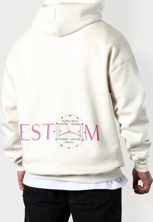 Person trägt einen cremefarbenen Hoodie mit pinker Schrift und einer Uhr-Grafik auf dem Rücken, kombiniert mit schwarzen Hosen und einem weißen Hemd darunter.