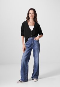 Schwarze gestrickte Strickjacke mit V-Ausschnitt und Knöpfen, kombiniert mit hoch taillierten, weiten blauen Jeans und schwarzen Sneakers.