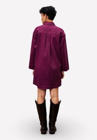 Bordeauxfarbendes Corduroy-Hemdkleid mit langen Ärmeln, einem Kragen und einem hinten eingearbeiteten Faltenzug. Schwarze knielange Stiefel runden das Outfit ab.