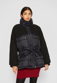 Svart pufferjakke med en teksturert mørk fleeceoverdel, frontknapper, elastisk livbånd og store klaftlommer.