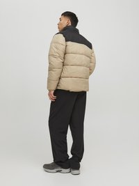 Giacca a vento con un corpo beige e spalle nere, caratterizzata da un colletto alto, texture trapuntata e vestibilità rilassata, indossata con pantaloni scuri.