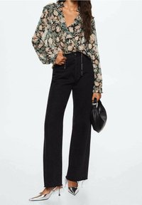 Femme portant un chemisier à volants fleuri rentré dans un jean large taille haute noir avec des ourlets effilochés, tenant un sac à main noir et portant des talons argentés.