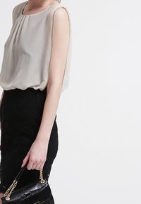 Femme portant un blouse sans manches beige et une jupe noire, tenant un sac à main en cuir noir avec une bandoulière en chaîne dorée devant un fond blanc.