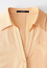 Camicia button-up color crema con colletto classico, texture morbida, tessuto leggero e un delicato luccichio, etichettata "un giorno".