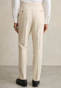 Pantalon beige clair à coupe ajustée, doté de plis doubles, de poches arrière avec fermeture à bouton et de chaussures habillées noires.