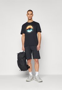 Sort t-shirt med farverig bjerg- og bølgedesign, parret med sorte shorts og grå vandresko, mens han/hun holder en sort rygsæk.