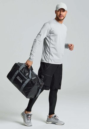 Chemise grise à manches longues, short noir, portant un sac de sport noir avec des accents gris et le mot "SLIM" imprimé. Chaussures de sport grises.