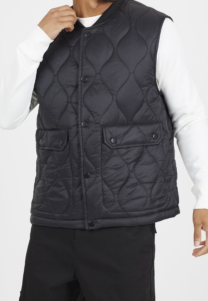 Black quilted vest with a rounded neckline, snap buttons, and two front pockets. Smooth fabric texture with a wavy pattern throughout.