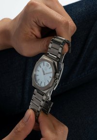 Mâini ținând un ceas de mână din metal argintiu cu un cadran bleu deschis și cifra romană XII, cu brățara desfăcută pe un fundal de țesătură închisă.