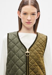 Γιλέκο καπιτονέ σε ελαιί πράσινο και χακί, με χρυσό φερμουάρ και στρογγυλεμένο λαιμόκοψη. Απαλό, γυαλιστερό ύφασμα με μοτίβο διαμαντιού.