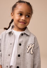 Chaqueta sudadera gris con botones negros, que presenta un bordado texturizado en crema con una "N" en el pecho izquierdo, usada sobre una camiseta blanca.