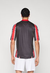 Lühikeste varrukatega must jersii, millel punakad ja valged triibulised aktsendid, ümara kaelusega ja sileda, kerge kangaga tekstuuriga.