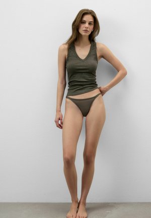 Ung kvinde stående barfodet mod en hvid væg iført en mørkegrøn ærmeløs top og matchende bikiniunderdele.