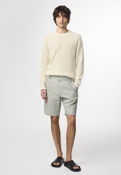 Cremefarbener, strukturiert gestrickter Pullover, hellgraue Shorts und schwarze Slidesandalen. Das Model steht vor einem neutralen Hintergrund und zeigt die Details des Outfits.