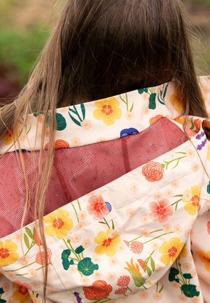 Langes braunes Haar, das über den Rücken eines Blumenregenmantels mit gelben, orangefarbenen und grünen Blumen fällt, der ein rotes Netzfutter zeigt.