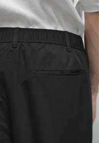 Pantaloni neri visti da dietro, con una fascia in vita e una piccola tasca aperta sul lato destro. Una camicia bianca è parzialmente visibile sopra.
