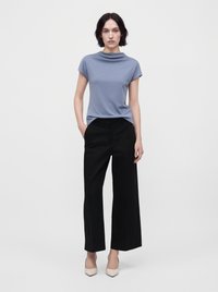 Femme aux cheveux courts et foncés, portant un haut bleu clair à manches courtes, un pantalon noir à jambes larges et des chaussures beiges pointues, debout sur fond blanc.