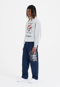 Lys grå sweatshirt med grafisk print; løse blå jeans med farverigt broderedesign; hvide sneakers.