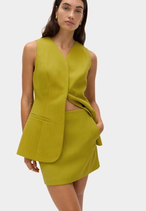 Vrouw die een mouwloze limegroene blazer en bijpassende minirok draagt, met één hand in de zak tegen een effen achtergrond.