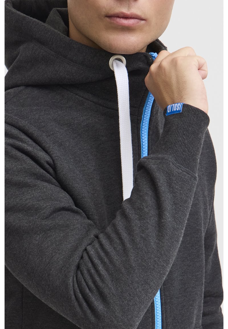 Donkergrijze hoodie met een blauwe rits, witte trekkoorden en geribde manchetten. Bevat een logo op de mouw. Zachte stofstructuur zichtbaar.