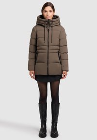 Frau, die eine braune gepolsterte Winterjacke mit hohem Kragen und Kapuze, einen schwarzen Rock, durchsichtige Strumpfhosen und schwarze kniehohe Stiefel trägt und steht.