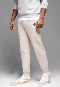 Béžové tepláky vyrobené z měkkého materiálu mají panelový design, žebrované manžety a černou cedulku na levém stehně. Doplněné o bílé tenisky.