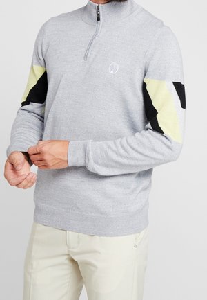Homme ajustant le poignet d'un pull gris clair à fermeture quart de zip avec des motifs géométriques noirs et jaunes sur les manches, portant un pantalon beige.
