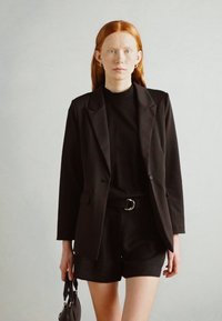 Black blazer over a black turtleneck, paired with black shorts. Model holds a small black handbag. Smooth fabric, tailored design.