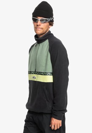 Pull en polaire noir et vert, doté d'une fermeture éclair demi-zip, d'une bande décorative à motifs et de poches latérales. Porté avec un bonnet noir et des lunettes de soleil.