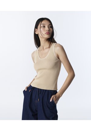 Giovane donna che indossa un top beige a coste senza maniche e pantaloni larghi blu scuro con le mani nelle tasche, che guarda di lato su uno sfondo bianco.