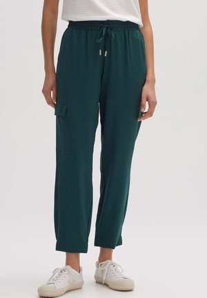 Persoon die donker groene cargobroek met trekkoord en omgeslagen pijpen draagt, samen met witte sneakers, staand tegen een effen achtergrond.