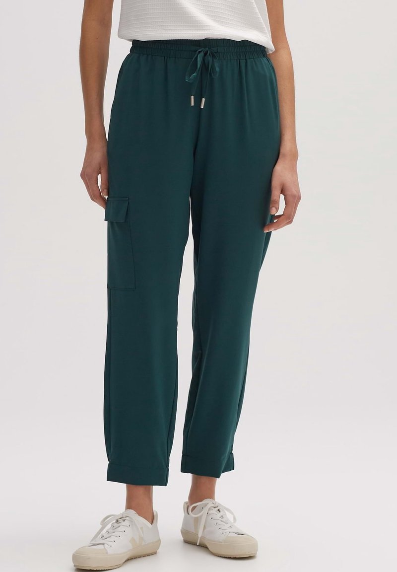 Personne portant un pantalon cargo vert foncé avec cordon de serrage et ourlets retroussés, ainsi que des baskets blanches, debout devant un fond uni.