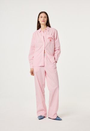 Vrouw draagt een pyjama met roze-witte verticale strepen en een geborduurd borstzakje, staat met één hand in haar zak en blauwe puntige schoenen.