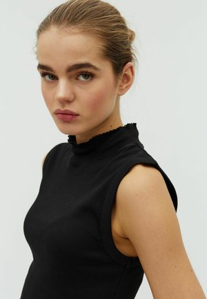 Ung kvinde med lyst brunt hår sat op i en knold, iført en ærmeløs sort top med høj, flæset halsudskæring, mod en ensartet lys baggrund.