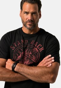 Camiseta de algodón negra con un estampado gráfico rojo que presenta utensilios cruzados y un diseño de llamas, mangas cortas, cuello redondo y corte relajado.