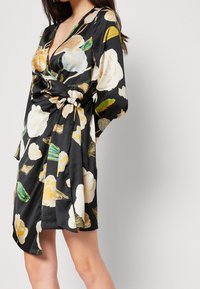 Robe enveloppante en satin noir ornée de grands motifs floraux en crème et jaune, avec des manches longues et un détail de taille nouée.
